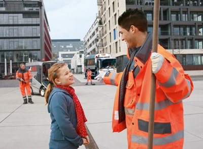 Stadtreiniger unterhält sich mit einem Mädchen in der Kölner Innenstadt. Im Hintergrund agieren zwei weitere Stadtreiniger mit Besen und eine AWB Kehrmaschine. 400x293px 