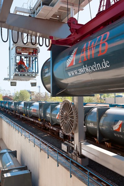 Müllumladestation mit Kranfahrer und einem AWB Container am Kran und einem Zug mit vielen AWB Containern auf der Schiene. 